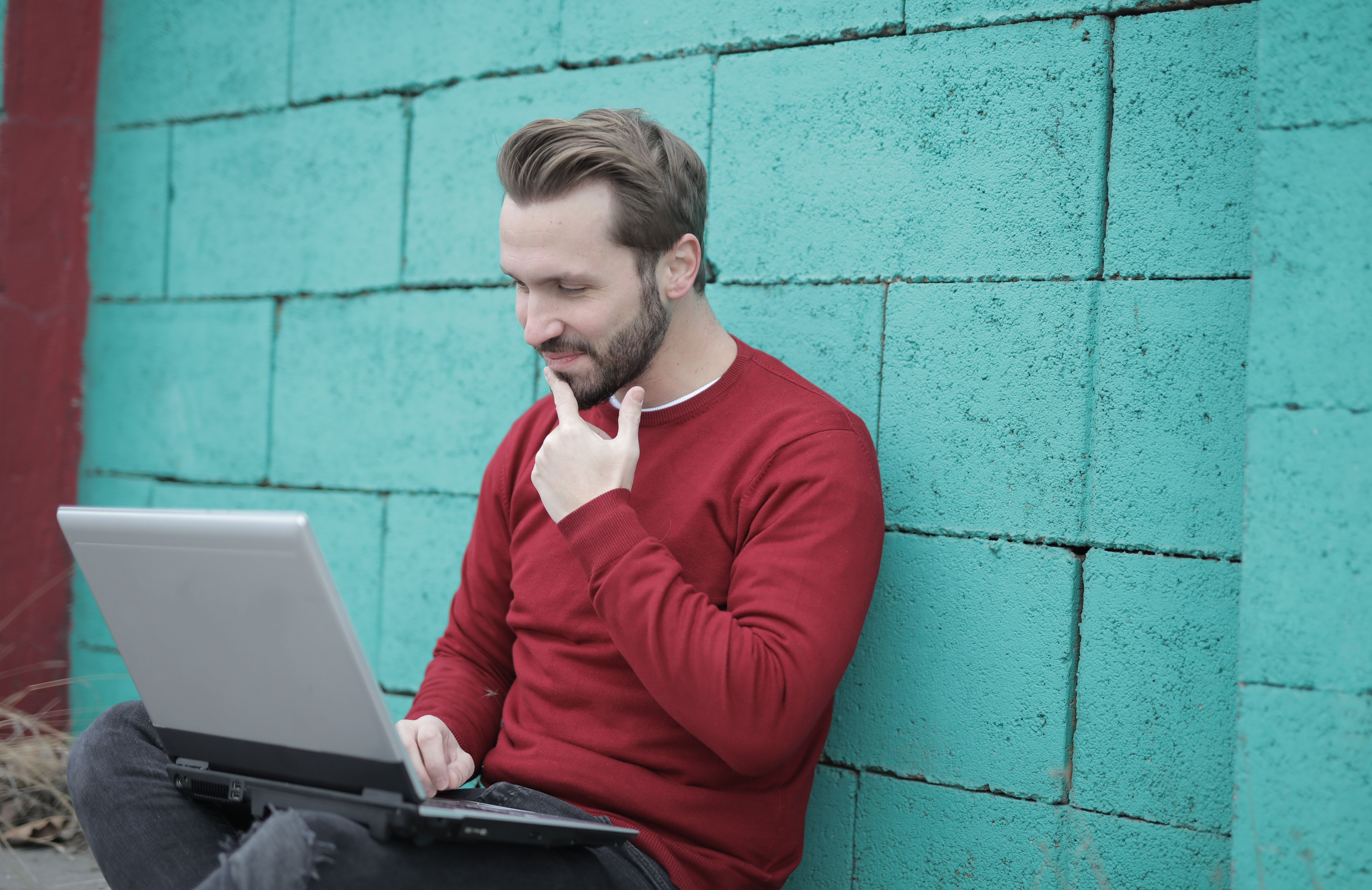 A man in a red sweater using a laptop against a turquoise wall.